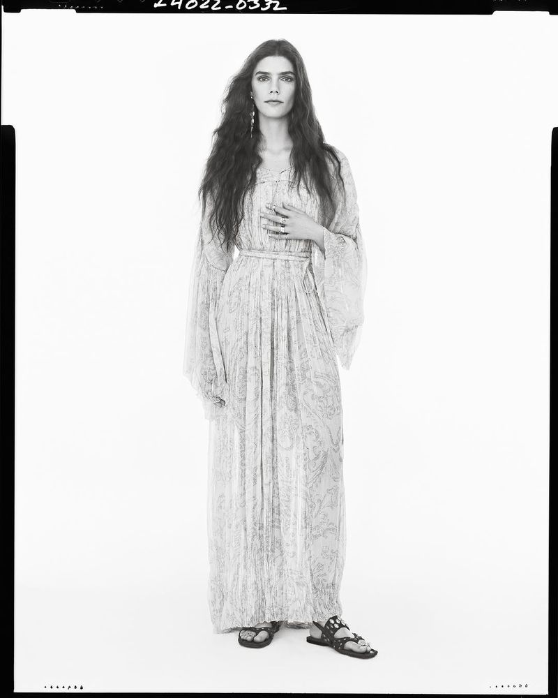 Full-length portrait of a female model with long, dark wavy hair, wearing a long-sleeved patterned dress, flat sandals, standing against a white background.