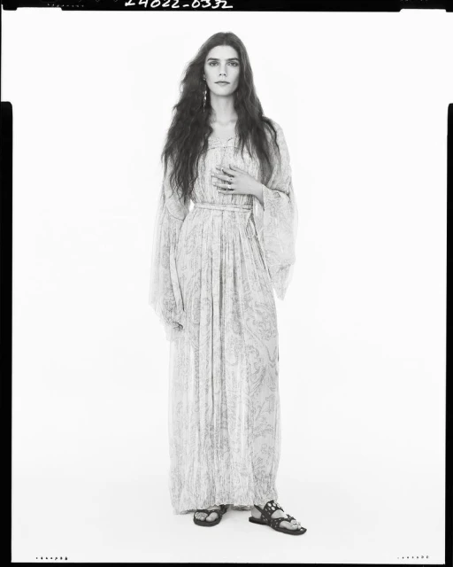 Full-length portrait of a female model with long, dark wavy hair, wearing a long-sleeved patterned dress, flat sandals, standing against a white background.