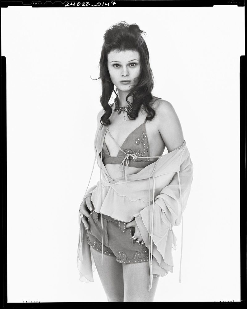 Monochrome portrait of a fair-skinned woman with curly dark hair, wearing a beaded top and shorts, and a sheer open blouse.