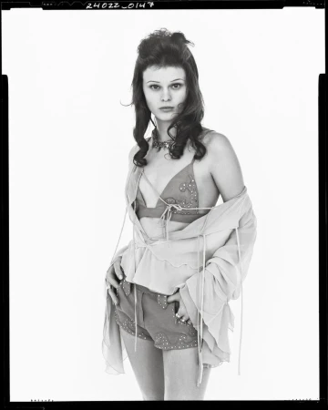 Monochrome portrait of a fair-skinned woman with curly dark hair, wearing a beaded top and shorts, and a sheer open blouse.
