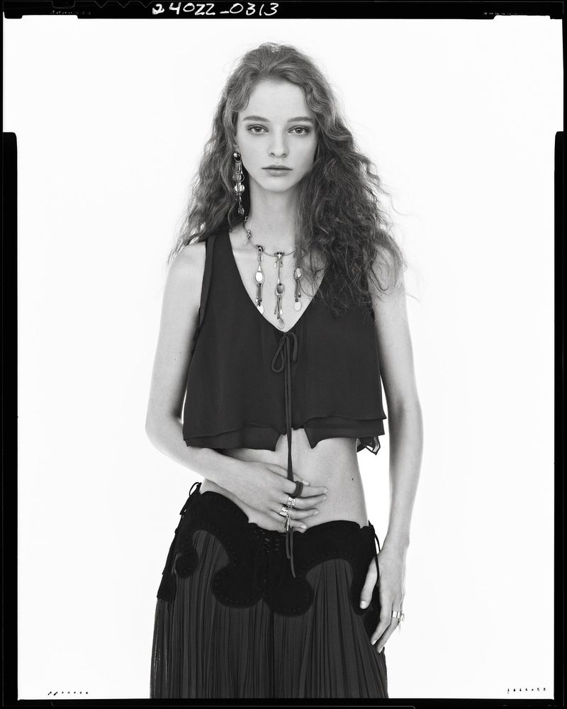 A slender woman poses in a black top and long black skirt against a bright background, accentuating her jewelry and toned midriff.