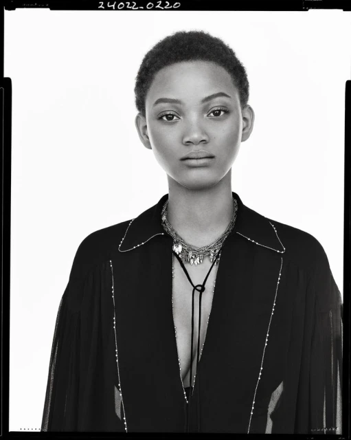 Headshot of a black female model with short hair, wearing a black top and jewelry, set against a bright white background in a studio.
