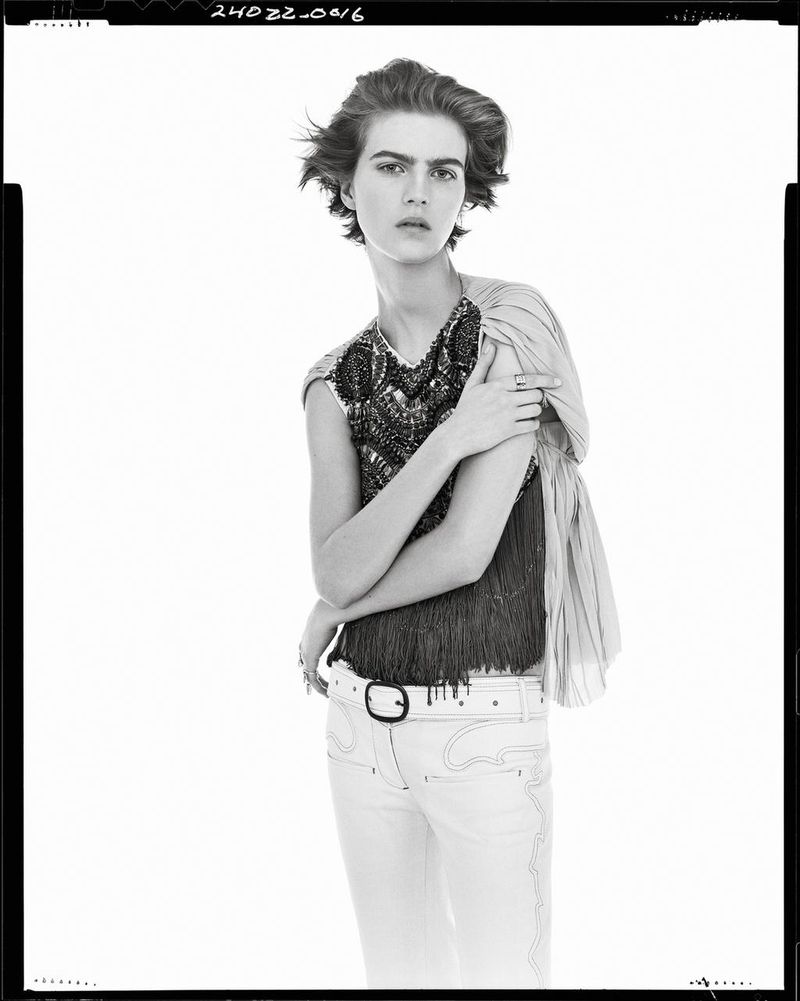 Model with short dark hair wearing a patterned sleeveless top and light trousers against white backdrop