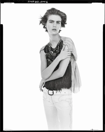 Model with short dark hair wearing a patterned sleeveless top and light trousers against white backdrop