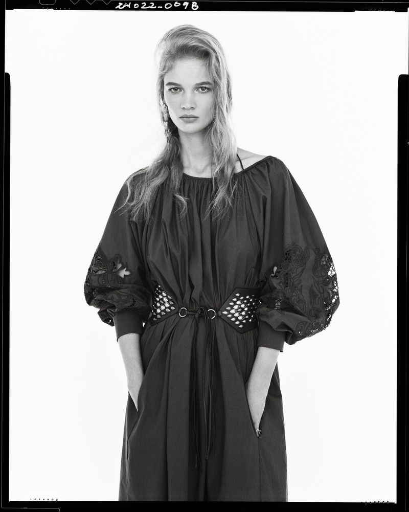 A fair-skinned woman with long blond hair poses indoors wearing a dark dress with embroidered sleeves and a studded belt against a white backdrop.