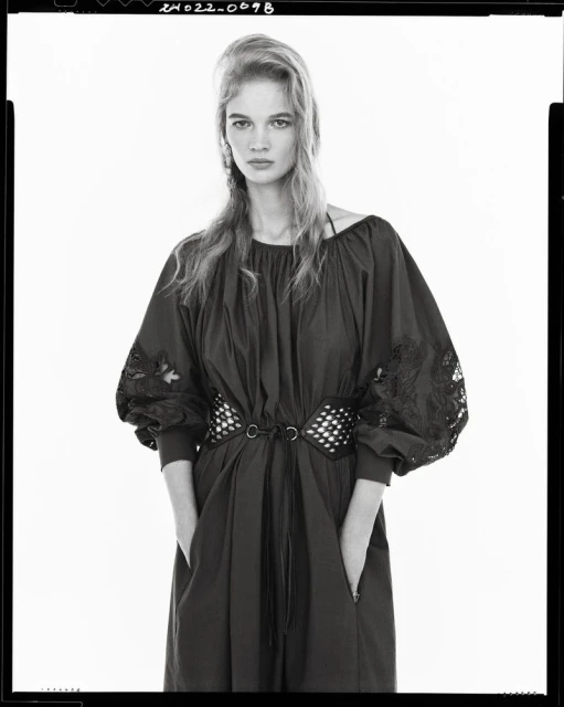 A fair-skinned woman with long blond hair poses indoors wearing a dark dress with embroidered sleeves and a studded belt against a white backdrop.