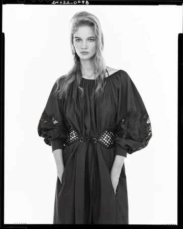 A fair-skinned woman with long blond hair poses indoors wearing a dark dress with embroidered sleeves and a studded belt against a white backdrop.