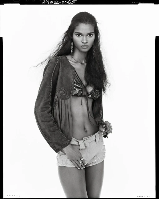 Fashion shot of a young adult woman with long dark hair, wearing a jacket, beaded bra top and beaded shorts, standing in a photo studio.