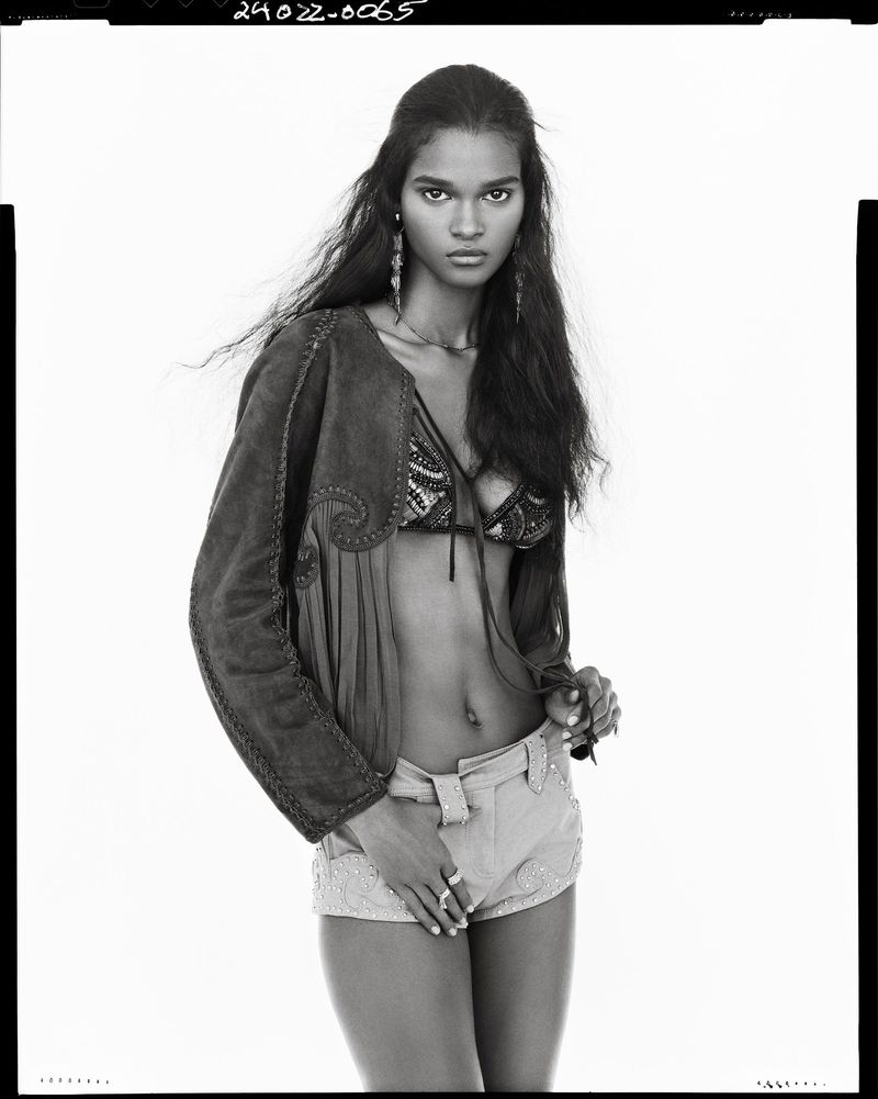 Black and white portrait of a model wearing a patterned jacket, bra top, and shorts against a white background