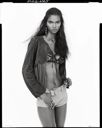 Black and white portrait of a model wearing a patterned jacket, bra top, and shorts against a white background
