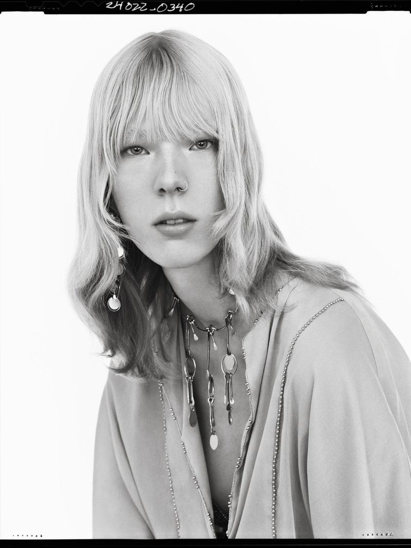 Black and white portrait of a model with blonde hair wearing layered necklaces against a white background
