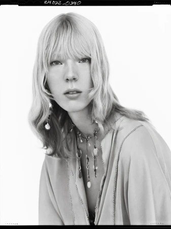 Black and white portrait of a model with blonde hair wearing layered necklaces against a white background