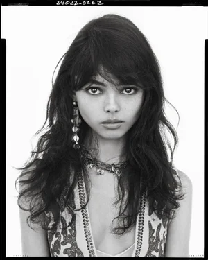 Black and white portrait of a woman with dark wavy hair wearing long statement earrings against a white background.
