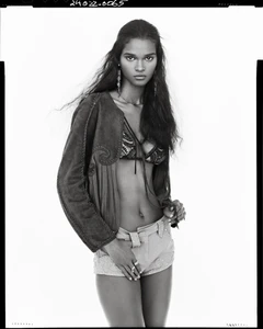 Black and white portrait of a model wearing a patterned jacket, bra top, and shorts against a white background