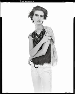 Model with short dark hair wearing a patterned sleeveless top and light trousers against white backdrop