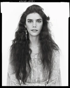 Portrait of a woman with long dark wavy hair wearing patterned apparel and long earrings against a white background