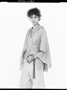 Model with short curly hair wearing a patterned tunic and layered bracelets against a white background