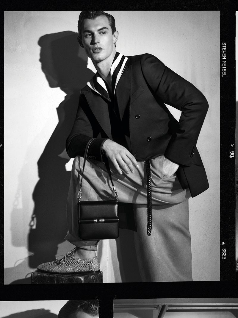 A man poses in a fashion shot in black and white, wearing a blazer, pants, and carrying a small bag. Shadow is visible in the background.
