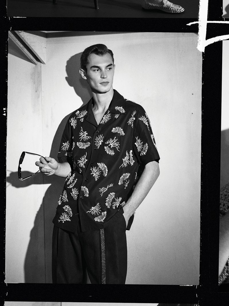 Model Kit Butler in a patterned black short-sleeve shirt holding sunglasses against a plain studio wall