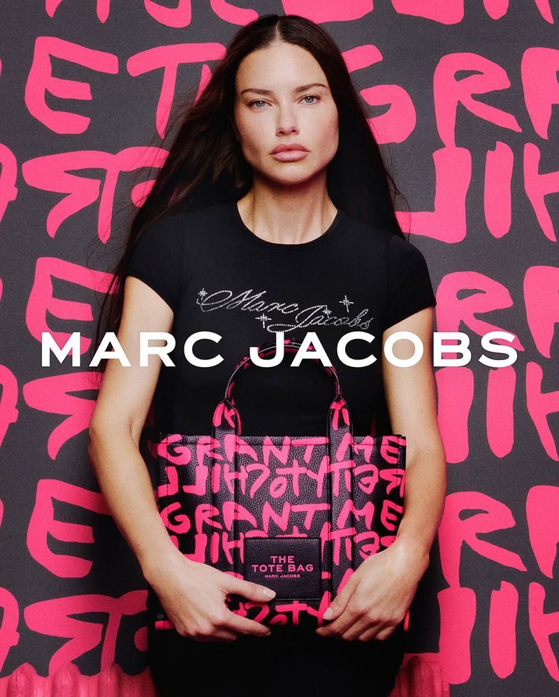 Woman poses in black Marc Jacobs t-shirt holding matching tote bag with pink graffiti text on a matching pink graffiti background.