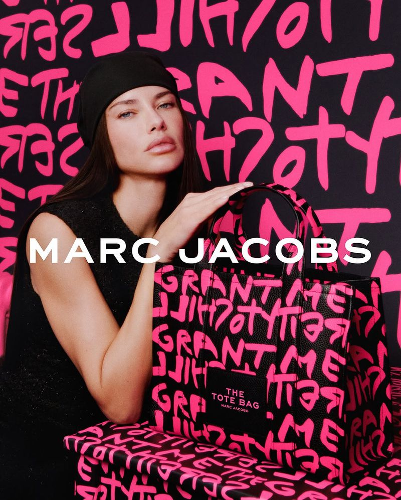 Fashion shot features a woman wearing a black beanie and dress, holding a Marc Jacobs tote bag with pink graffiti-style text, indoors.