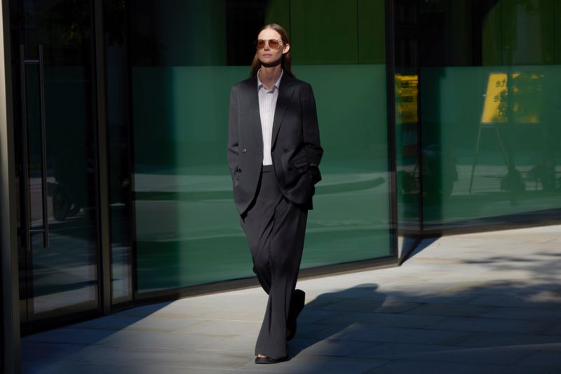 A woman in a dark suit walks in an outdoor urban setting, illuminated by sunlight and shadows, next to a modern glass building.
