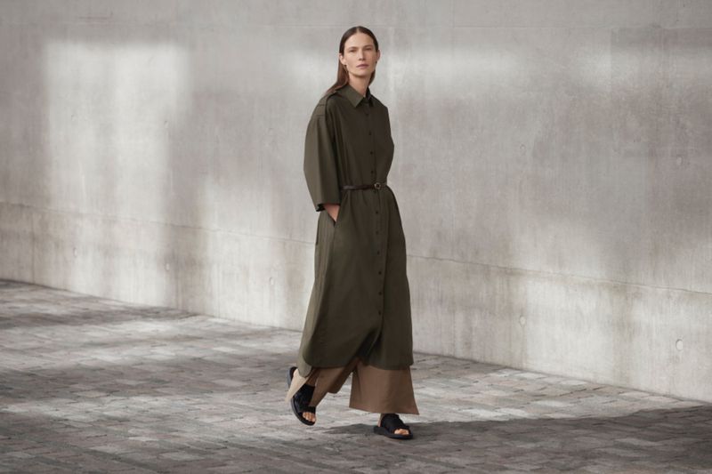 Full-length shot of a fashion model posing outdoors, wearing an olive-green belted dress and black sandals, against a concrete wall.