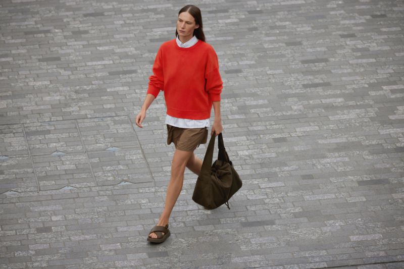 Fashionable adult female strides across grey brick wearing a red crewneck sweater, brown shorts, sandals, and carrying a slouchy bag.