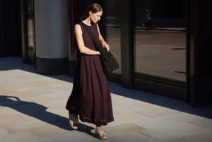 Model in a long dark dress and strappy sandals walking down a paved street while carrying a black shoulder bag.