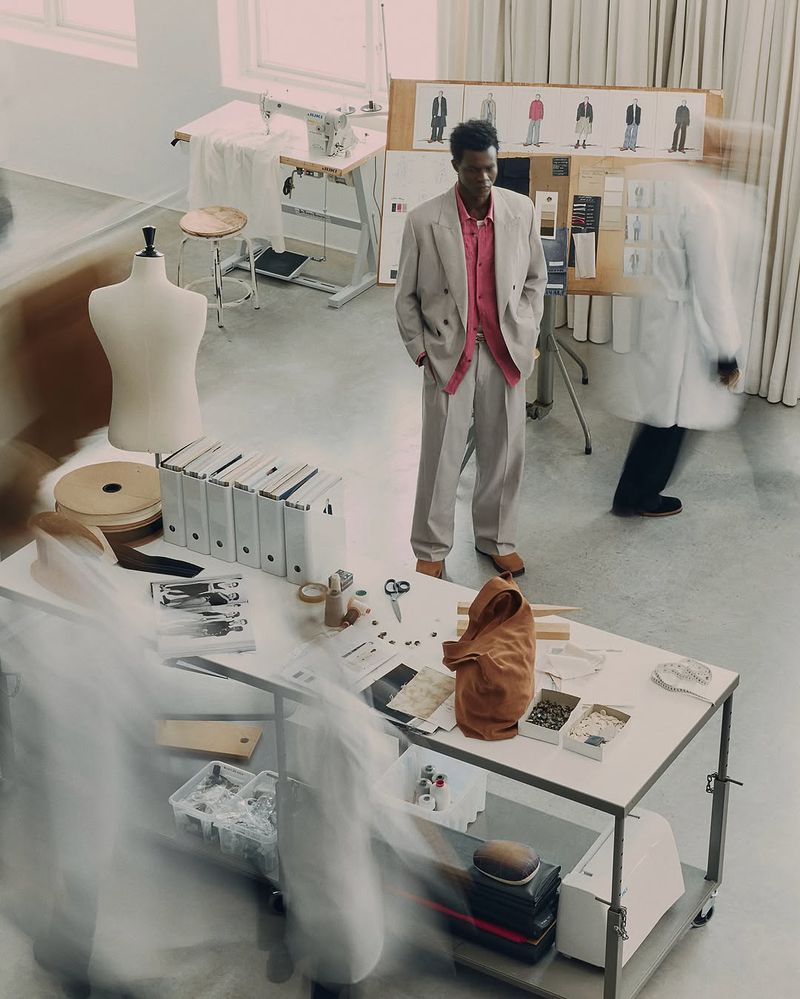 Fashion shoot of an adult Black male in a suit in a sewing studio with sewing machine, dress form, fabric swatches, and design sketches.