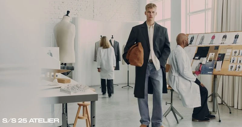 Fashion shoot in an atelier featuring a male model in a coat and jeans, posing with a leather bag, with design sketches in the background.