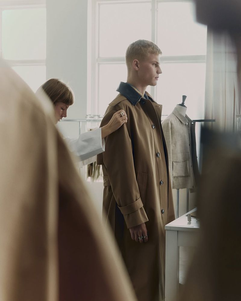 A male fashion model is standing indoors while a designer adjusts a brown trench coat in a bright photo studio setting.