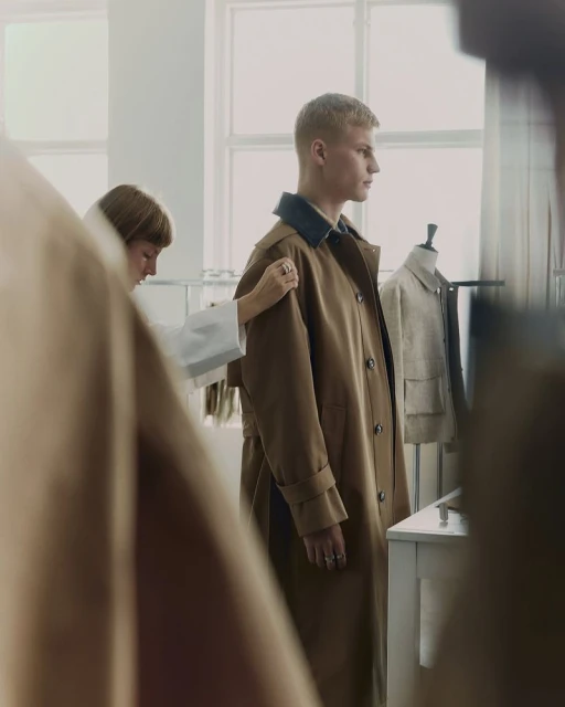 A male fashion model is standing indoors while a designer adjusts a brown trench coat in a bright photo studio setting.