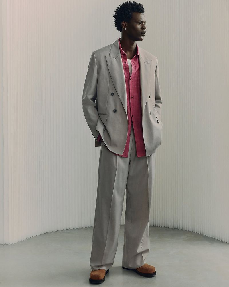 Full-length portrait of a male model wearing a grey double-breasted suit and a pink shirt against a neutral textured background.