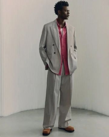 Full-length portrait of a male model wearing a grey double-breasted suit and a pink shirt against a neutral textured background.