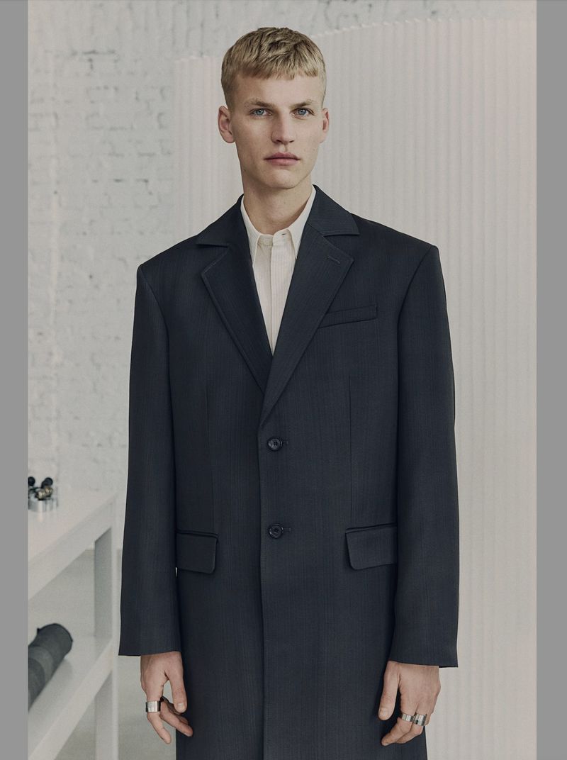 A young adult male wearing a dark overcoat and light collared shirt poses indoors against a minimalist white wall with shelf and camera.
