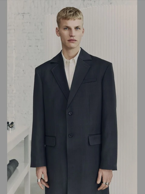 A young adult male wearing a dark overcoat and light collared shirt poses indoors against a minimalist white wall with shelf and camera.