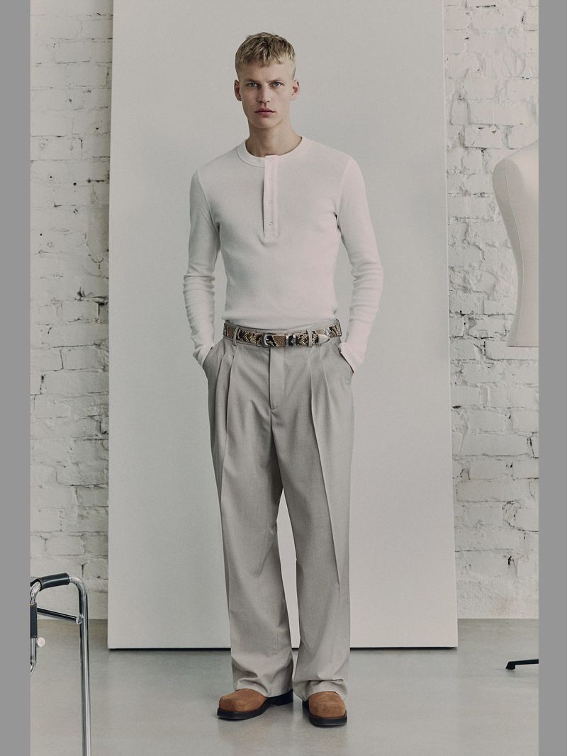 A full studio shot of a young adult male model in an off-white, long-sleeved henley shirt and light gray dress pants with an ornate belt.