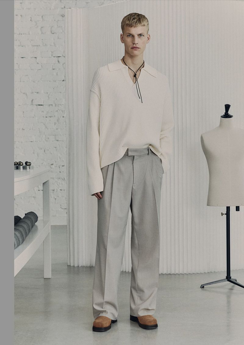 Model wearing a cream polo sweater and grey trousers standing in a studio