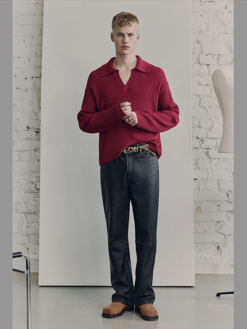 A fair-skinned adult male stands in a brightly lit studio. He's wearing a burgundy ribbed polo sweater, black leather pants, and brown suede shoes.