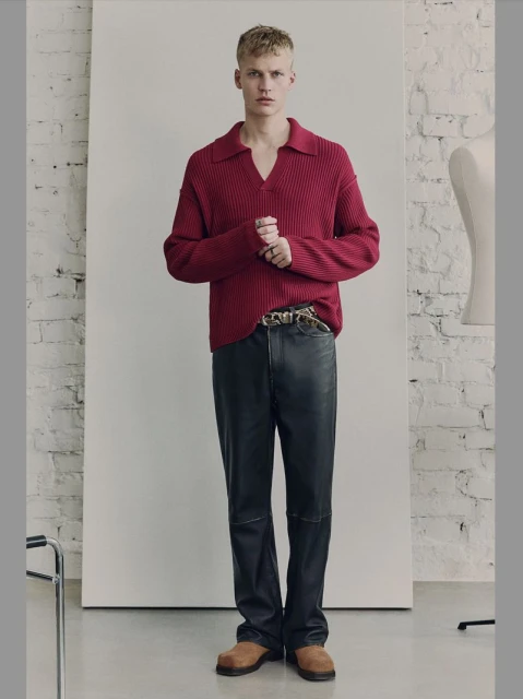 A fair-skinned adult male stands in a brightly lit studio. He's wearing a burgundy ribbed polo sweater, black leather pants, and brown suede shoes.
