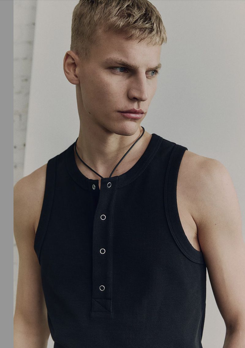 Close-up of a blond adult male in a black tank top with buttons, posing in front of a white wall in a brightly lit indoor environment.