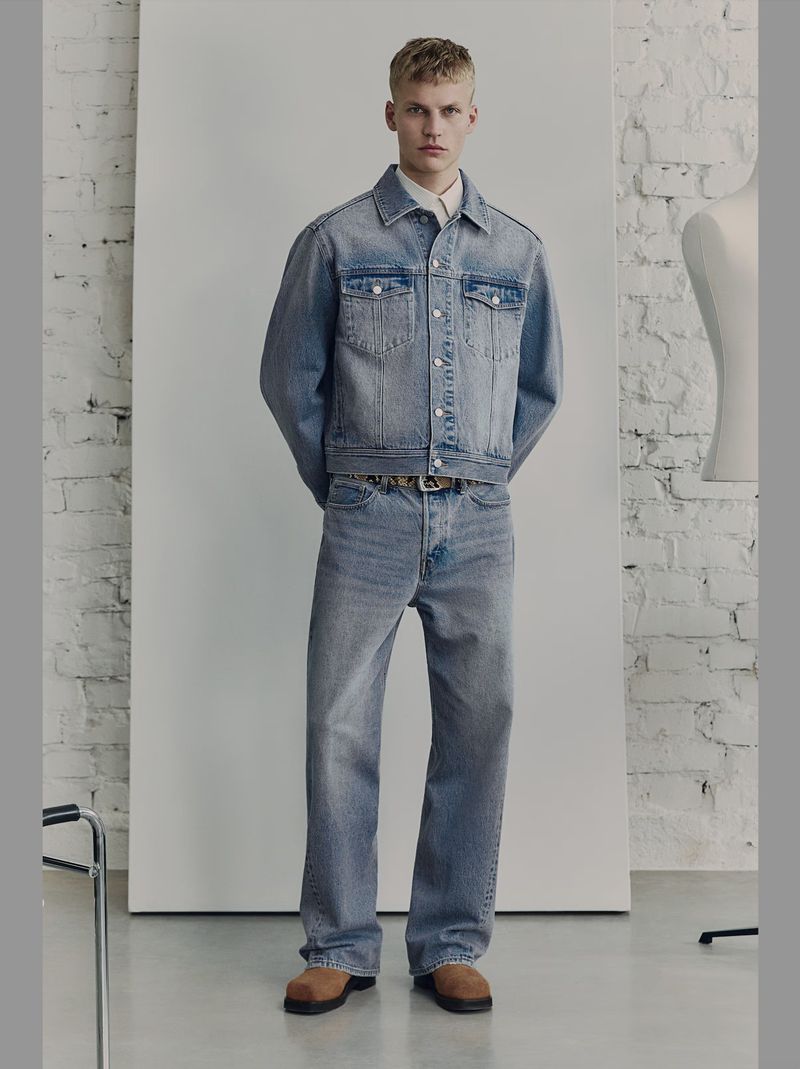 Full-length shot of a young adult male model wearing a denim jacket, jeans, button-down shirt, and belt posing indoors against a neutral backdrop.