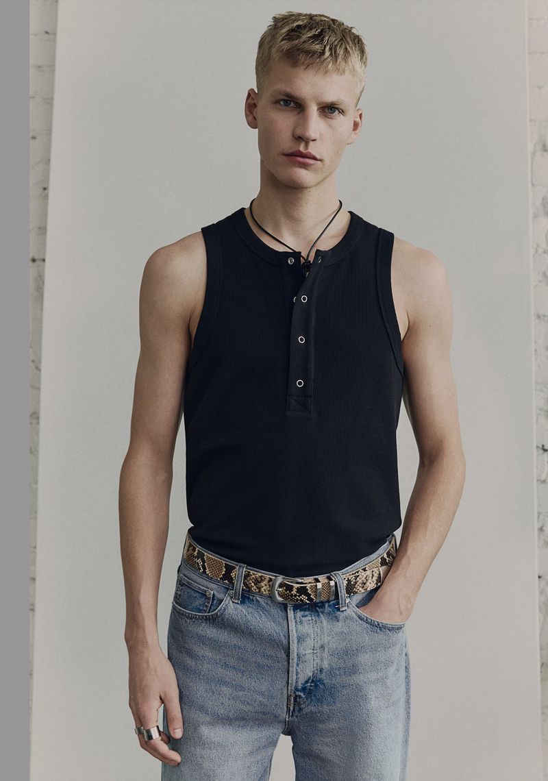 A blond male model poses in a black tank top, jeans, and a snakeskin belt, standing in front of a white wall in a studio setting.