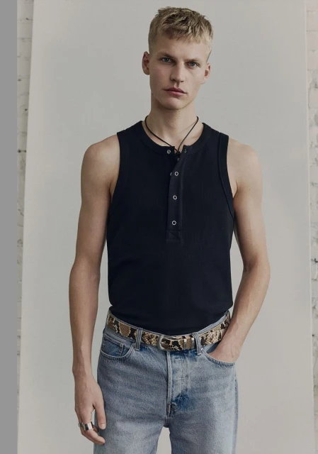 A blond male model poses in a black tank top, jeans, and a snakeskin belt, standing in front of a white wall in a studio setting.