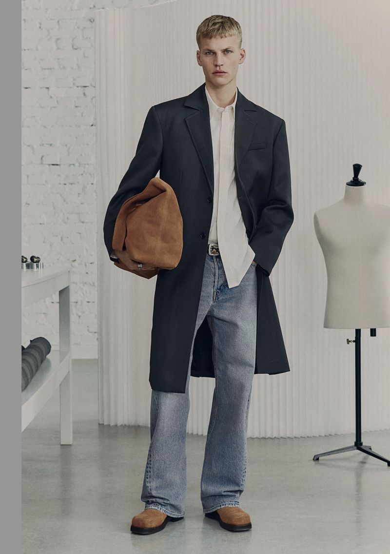 Male model wears a dark coat, light shirt, jeans, and carries a tan bag in a fashion editorial shot in an indoor studio setting.