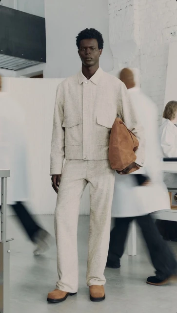 Dark-skinned male model wearing a light beige suit and brown leather shoes in a blurred interior setting with other people.