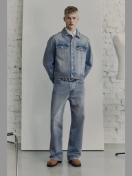 Full-length portrait of a male model wearing a denim jacket and jeans standing in a studio with a white backdrop and exposed brick wall.