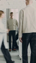 Model wearing a light jacket and dark trousers standing before a tall mirror against a white brick wall.