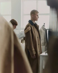 Behind-the-scenes fitting with a blonde male model wearing a brown trench coat in a studio setting